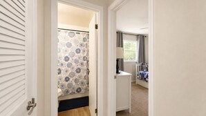 Een gang die leidt naar een badkamer met een douchegordijn met een patroon, een nachtkastje met een lamp en een bed met een quilt.