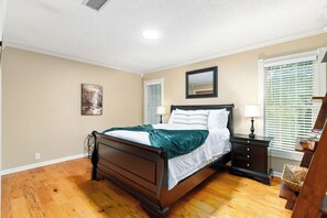 Une chambre à coucher comprenant un lit, des tables de chevet, une commode, un miroir et une fenêtre avec des stores.