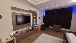 Quarto de hotel moderno com televisão de tela plana, escrivaninha com cadeira, sofá e uma janela grande com cortinas.