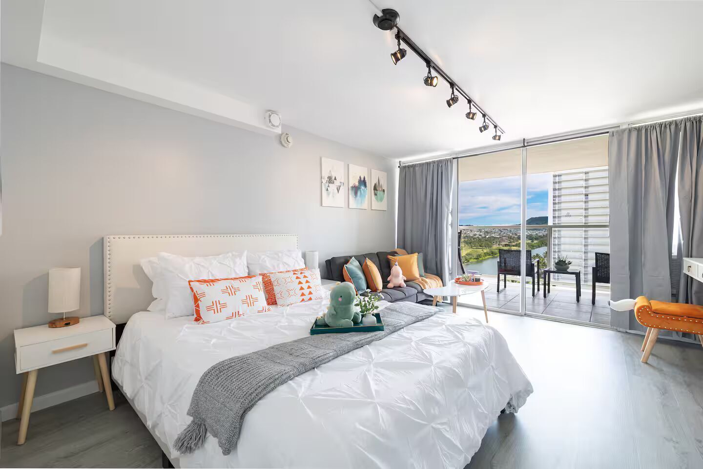 A modern bedroom with a large bed, a nightstand, a bedside table, a sofa, a small table, and a view of the outdoors through a sliding glass door.