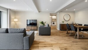 A modern living room with a flat-screen TV, grey sofas, and a dining area with a wooden table and chairs. - Oceanea Apartment by Homie (Santa Cruz)