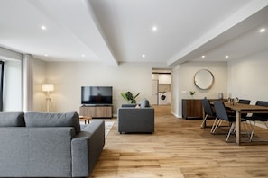 Ein modernes Wohnzimmer mit Flachbildfernseher, grauen Sofas und einem Essbereich mit Holztisch und Stühlen.