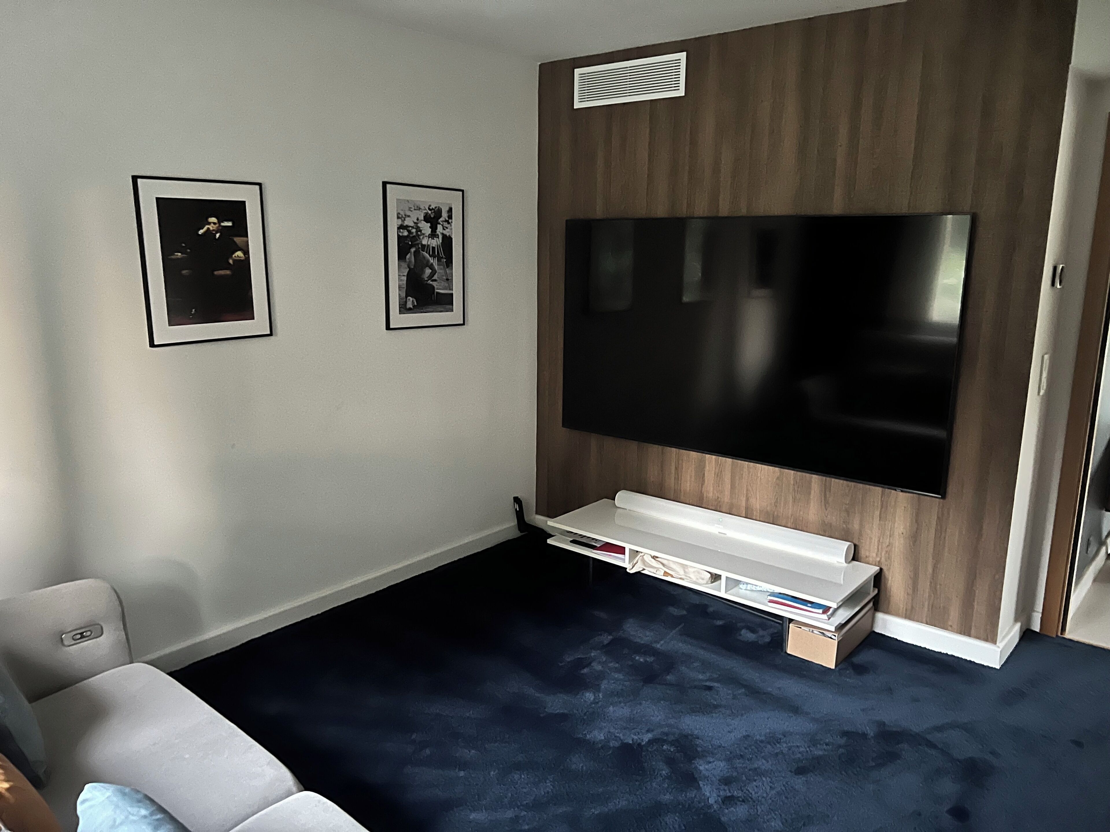 Quarto com uma televisão de tela grande fixada numa parede de madeira, um móvel branco embaixo e duas molduras na parede.