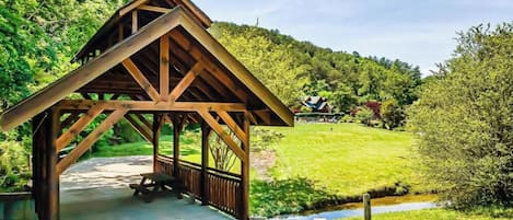 Um pavilhão de madeira com um banco, rodeado por vegetação exuberante e um pequeno riacho.