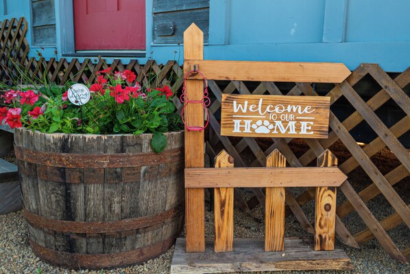A wooden sign with 'Welcome to Our Home' text, a rustic barrel, and a wooden fence with flowers.