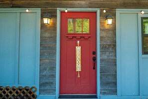 Une porte rouge avec une pancarte « BIENVENUE », un revêtement en bois et deux lampes fixées au mur.