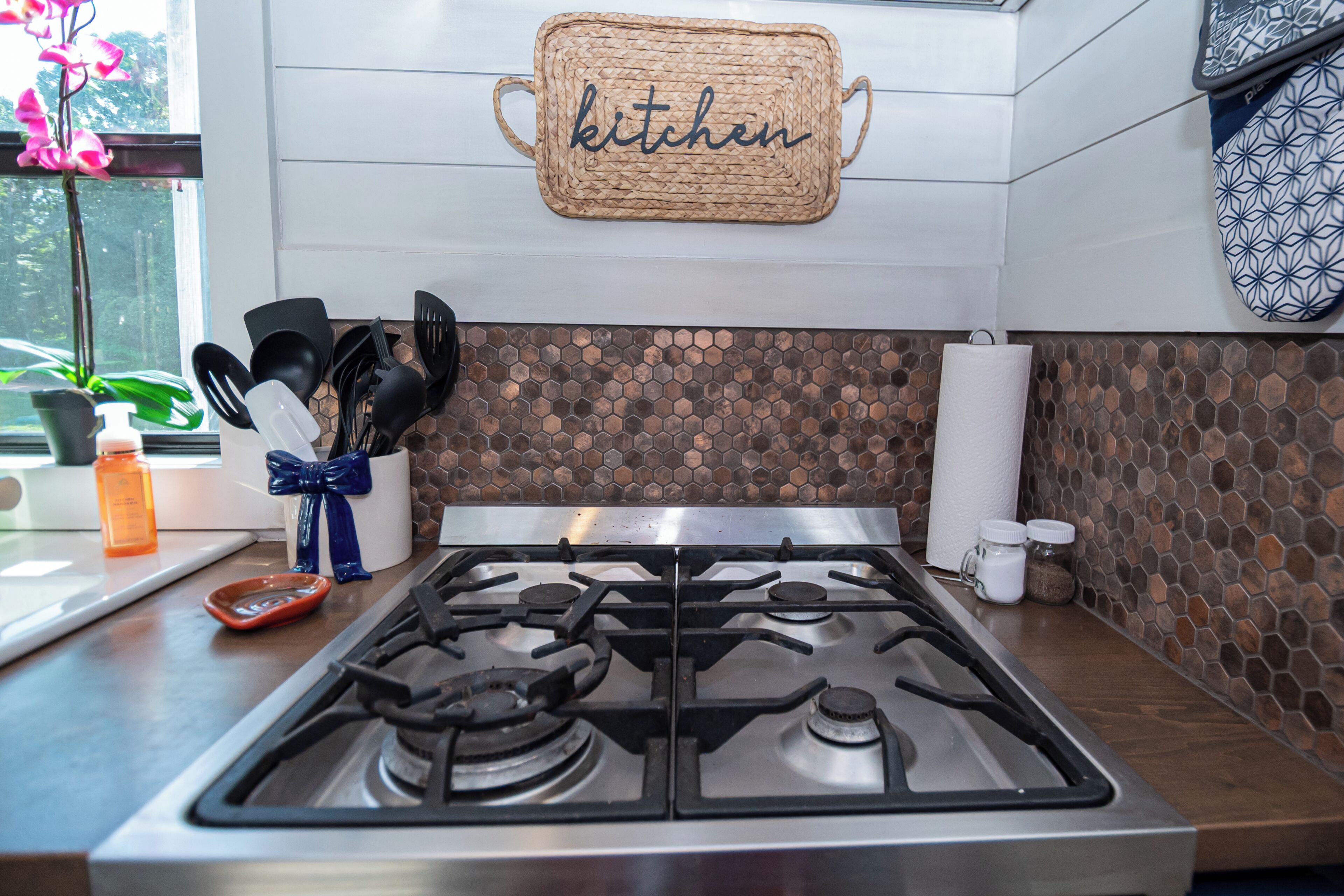 A kitchen with a gas stove, a woven kitchen sign, and a hexagonal tile backsplash.