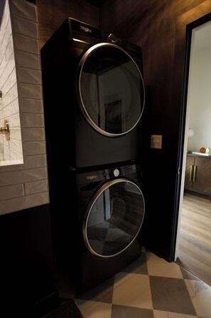 Una unidad de lavadora y secadora Whirlpool negra con panel de control, ubicada en una habitación con piso de cuadros y pared de azulejos blancos.