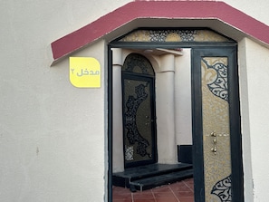 Ein Gebäudeeingang mit einer verzierten Tür, einem gelben Schild mit arabischer Schrift und einem roten Dach.