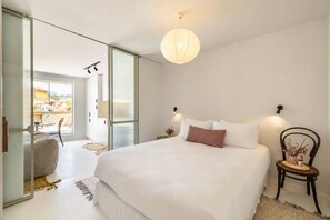 Ein Schlafzimmer mit einem großen Bett, einem Sessel, einem kleinen Tisch und Blick durch eine Glastür auf einen Außenbereich.
