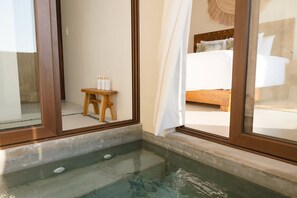 Une pièce dotée d’un jacuzzi entouré de parois en verre, d’un tabouret en bois sur lequel sont posés des articles de toilette, et d’un lit avec une tête de lit.