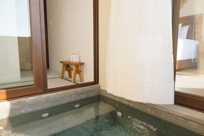A corner of a room with a small wooden stool and bottles on a table, a bathtub, and a window with a view of a bed. - Studio Villa at Cerro del Vigia (Zihuatanejo)
