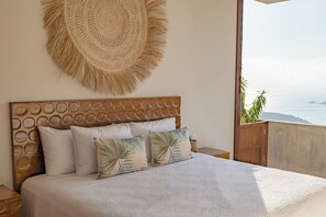 Een slaapkamer met een bed, kussens, een hoofdbord, een wanddecoratie en uitzicht op zee.