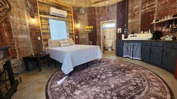 A rustic room with a bed, a round rug, a wooden ceiling, and a kitchen area.