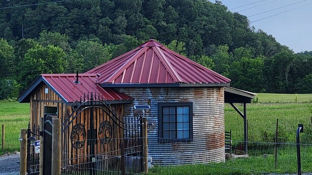 Een rustieke houten chalet met een metalen dak, omgeven door een hek en een omheining, gelegen voor een groene heuvel.