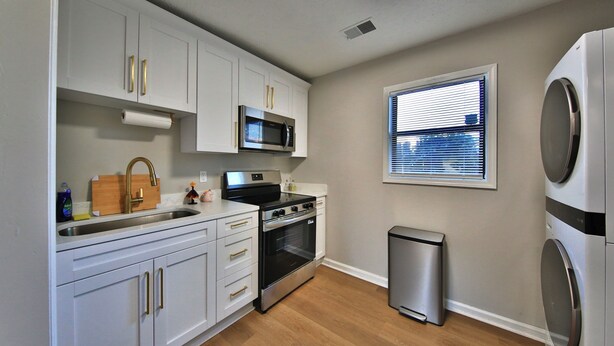 A compact kitchen with white cabinets, a stainless steel microwave, an oven, a sink, and a small trash bin.