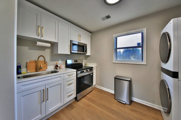 A compact kitchen with white cabinets, a stainless steel microwave, an oven, a sink, and a small trash bin.