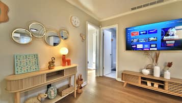 Un salón con un televisor de pantalla plana montado en la pared, una mesa consola de madera con objetos decorativos, y un pasillo que conduce a otra habitación.