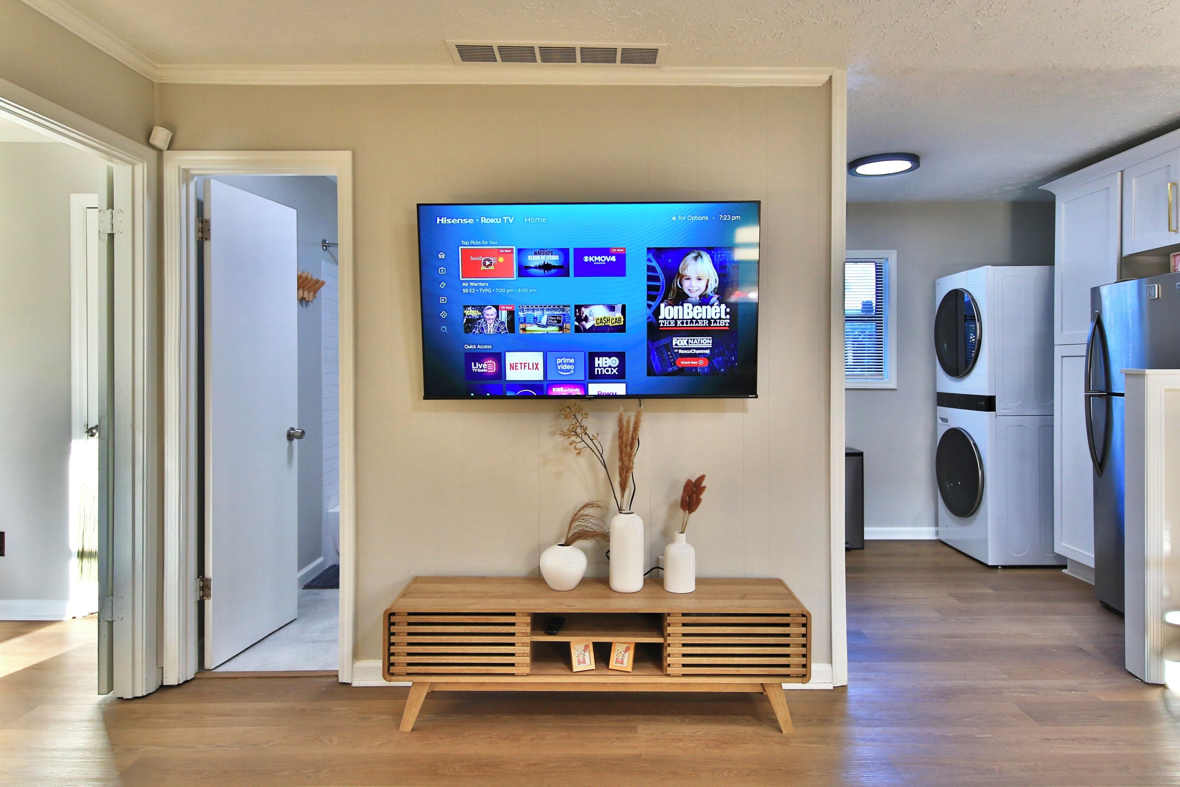 A modern living room with a flat-screen TV mounted on the wall, a wooden console table with decorative vases, and a laundry area with a washing machine and dryer.