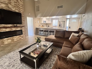 A modern living room with a sectional sofa, a glass coffee table, and a flat-screen TV mounted on a stone wall. - Luxury Living in Palm Desert! 2 Bed/2 Bath with a West Facing Patio (Palm Desert)