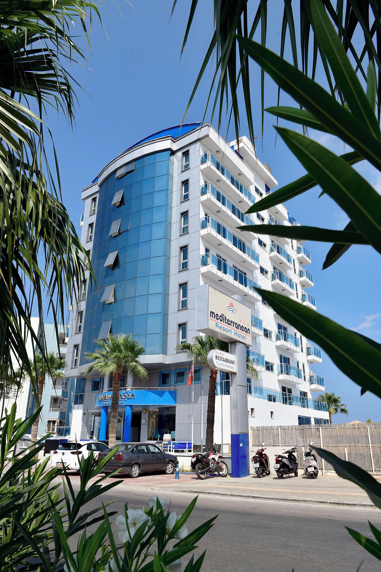 Un hôtel à plusieurs étages, doté d’une façade bleue et blanche, d’un restaurant et d’un parking où sont stationnées des voitures et des motos.