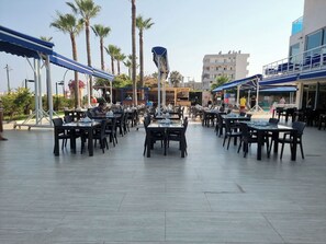 An outdoor dining area with multiple tables and chairs, surrounded by palm trees and buildings. - Mediterranean Resort Hotel (Silifke)