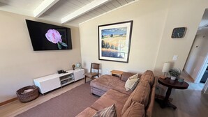A living room with a large brown sectional sofa, a white entertainment unit, a wall-mounted TV, and a framed landscape painting.
