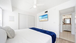 A bedroom with a bed, a ceiling fan, a TV, and a view of the bathroom.