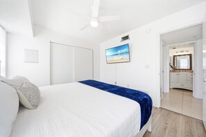 A bedroom with a bed, a ceiling fan, a TV, and a view of the bathroom.