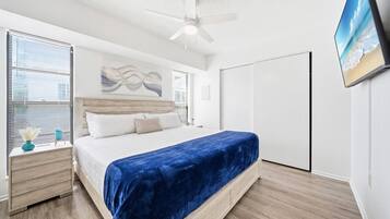 Une chambre à coucher comprenant un lit, des tables de chevet, un ventilateur de plafond, une grande fenêtre avec des stores et un téléviseur fixé au mur.