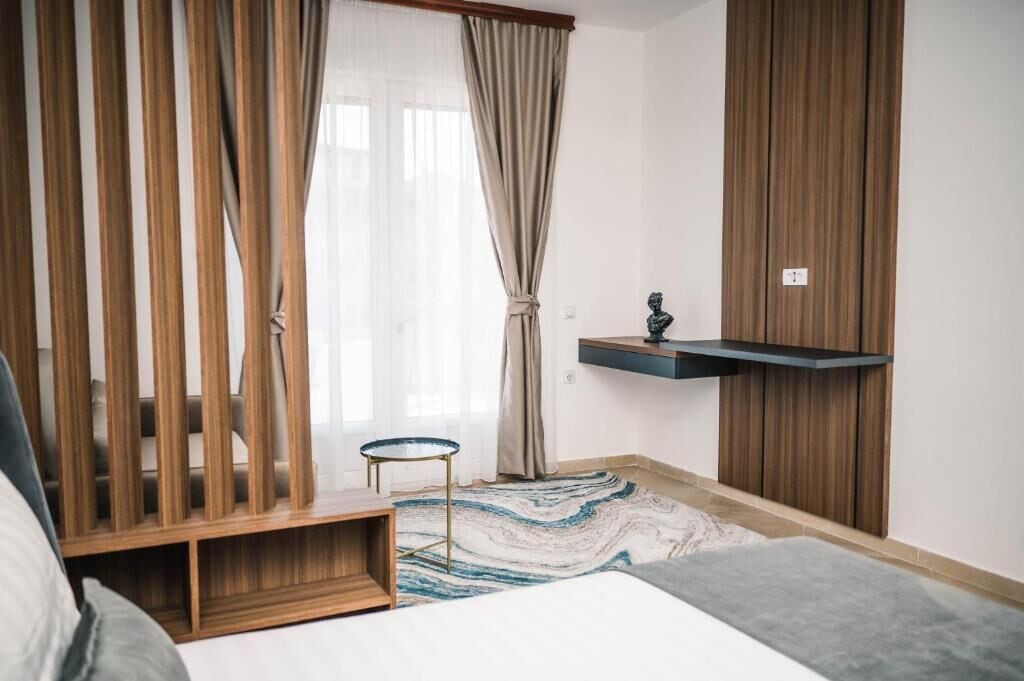 Een slaapkamer met een bed, een klein rond tafeltje, een houten kast, een hangplank en een groot raam met gordijnen.