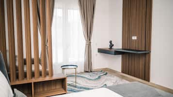 Een slaapkamer met een bed, een klein rond tafeltje, een houten kast, een hangplank en een groot raam met gordijnen.