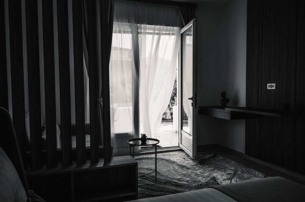 Una habitación con cama, una mesita, una ventana con cortinas de gasa y vistas al exterior.