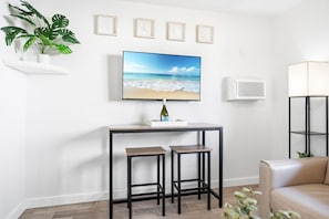 Ein modernes Wohnzimmer mit einem Flachbildfernseher, einem kleinen Tisch mit zwei Hocker, einer Topfpflanze und einem beigefarbenen Sofa.