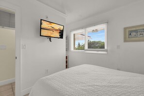 Ein Schlafzimmer mit einem Bett, einem an der Wand befestigten Fernseher, einem Fenster mit Jalousien und einer Tür.