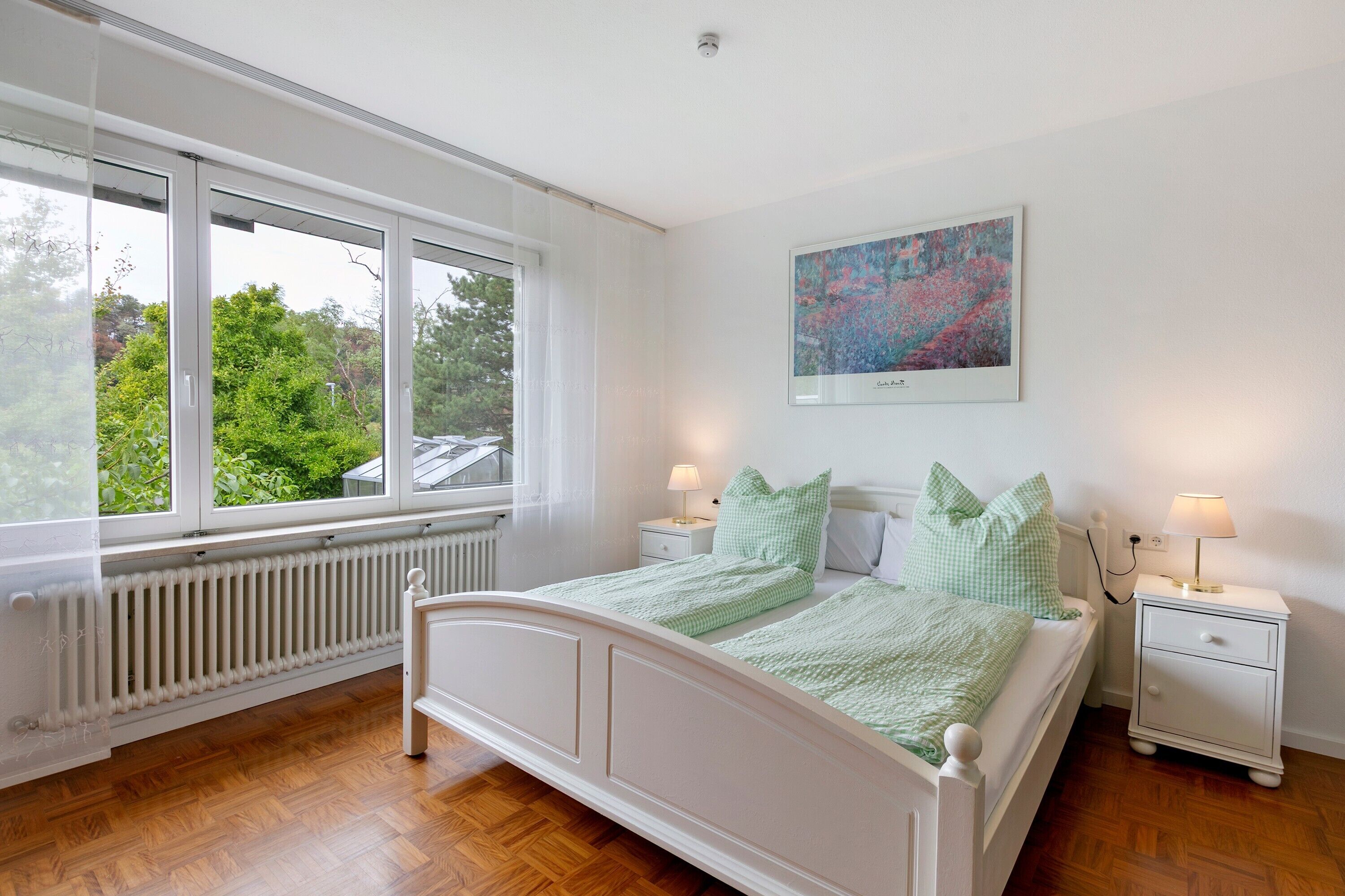 Ein Schlafzimmer mit einem Bett, zwei Nachttischen, einem Heizkörper, einem Fenster und einem Bild an der Wand.