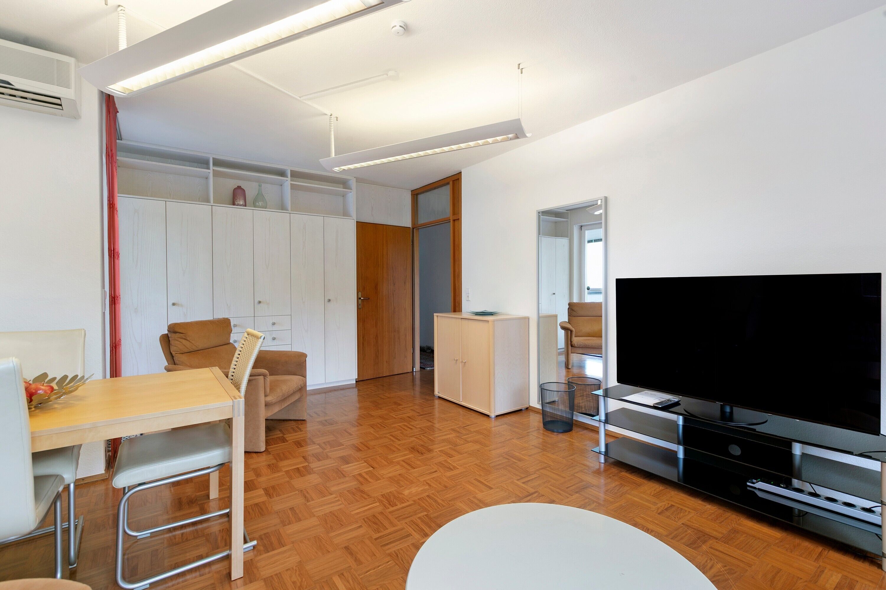 Una sala moderna con televisor de pantalla plana, piso de madera, una mesa de comedor con sillas, un sofá y un armario.
