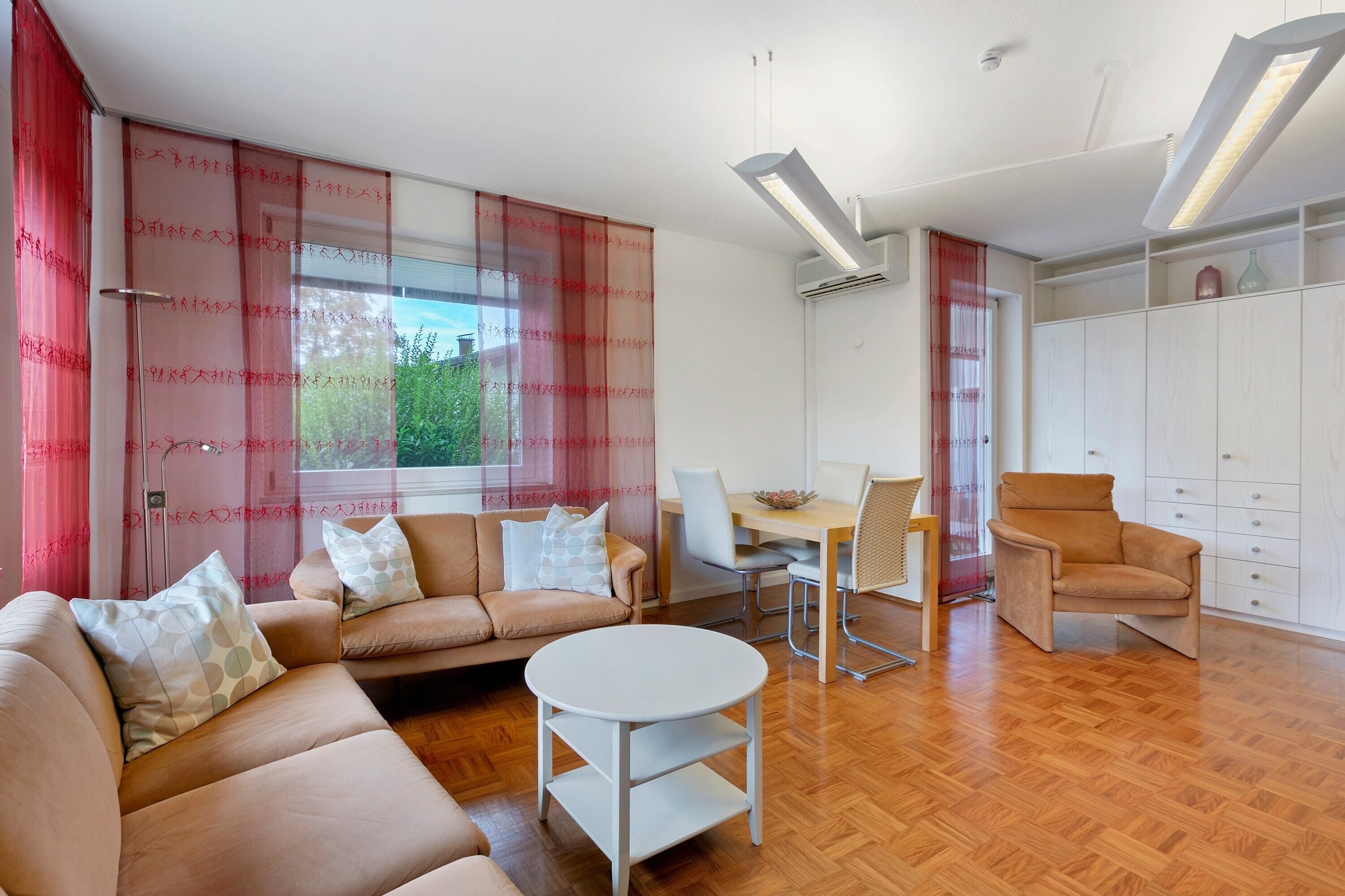 Ein modernes Wohnzimmer mit einer Sofagarnitur, einem Esstisch mit Stühlen, einem kleinen runden Tisch und einem Kleiderschrank.