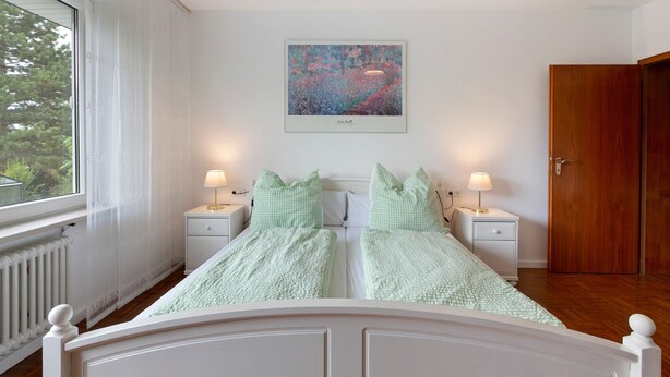 A bedroom with a double bed, two bedside tables with lamps, a window with curtains, a door, and a painting on the wall. - Quiet Riverside Wallbach Apartment with Terrace, AC, Washer, and Rhine View (Bad Säckingen)