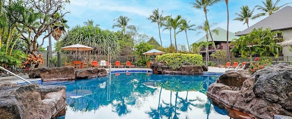 Zona de piscina con sillas de descanso y sombrillas, rodeada de vegetación tropical y un edificio con techo de paja.