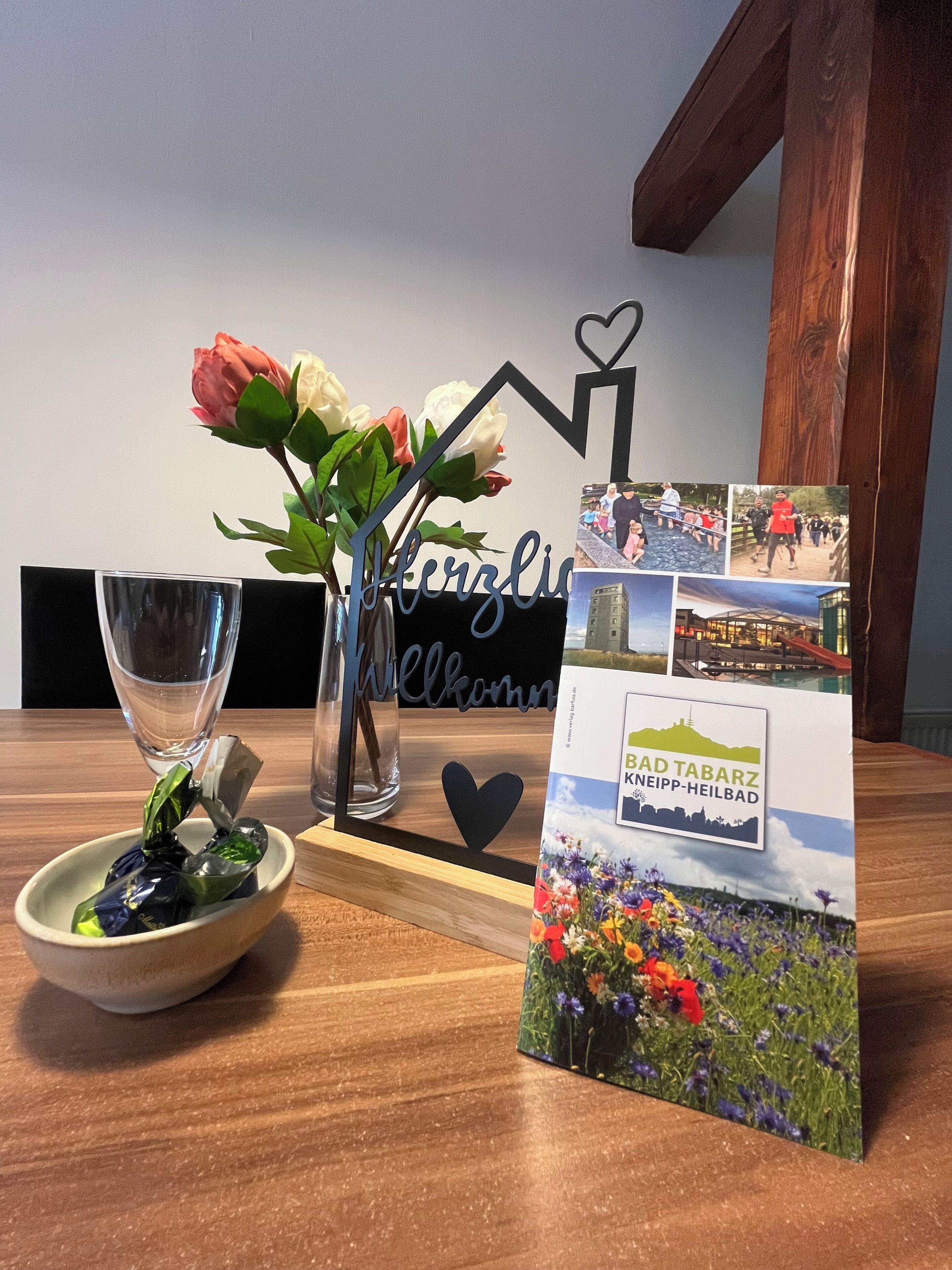 Una mesa de madera con un florero de vidrio con flores, un tazón de bombones, un vaso de agua y un cartel de bienvenida con un recorte en forma de corazón.