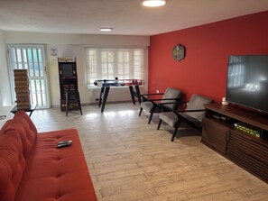 A living room with a red wall, a television, a sofa, a table with a foosball table, and a bookshelf.