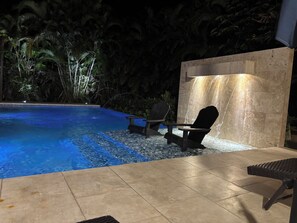 An outdoor swimming pool with a waterfall feature, surrounded by tropical plants and furnished with lounge chairs.