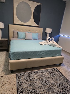 A bedroom with a bed, pillows, a nightstand, a lamp, a wall art piece, and a patterned rug.