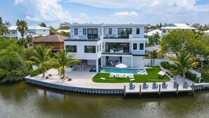 Une maison moderne à deux étages avec une piscine, entourée de palmiers et dotée d’un quai.