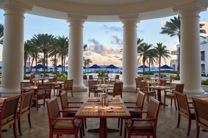 A covered outdoor dining area with tables and chairs, palm trees, and a view of a pool and building in the background. - Outstanding apartment at Dawn Beach Club, JW Marriott Resort, Beach and Spa (oyster pond)