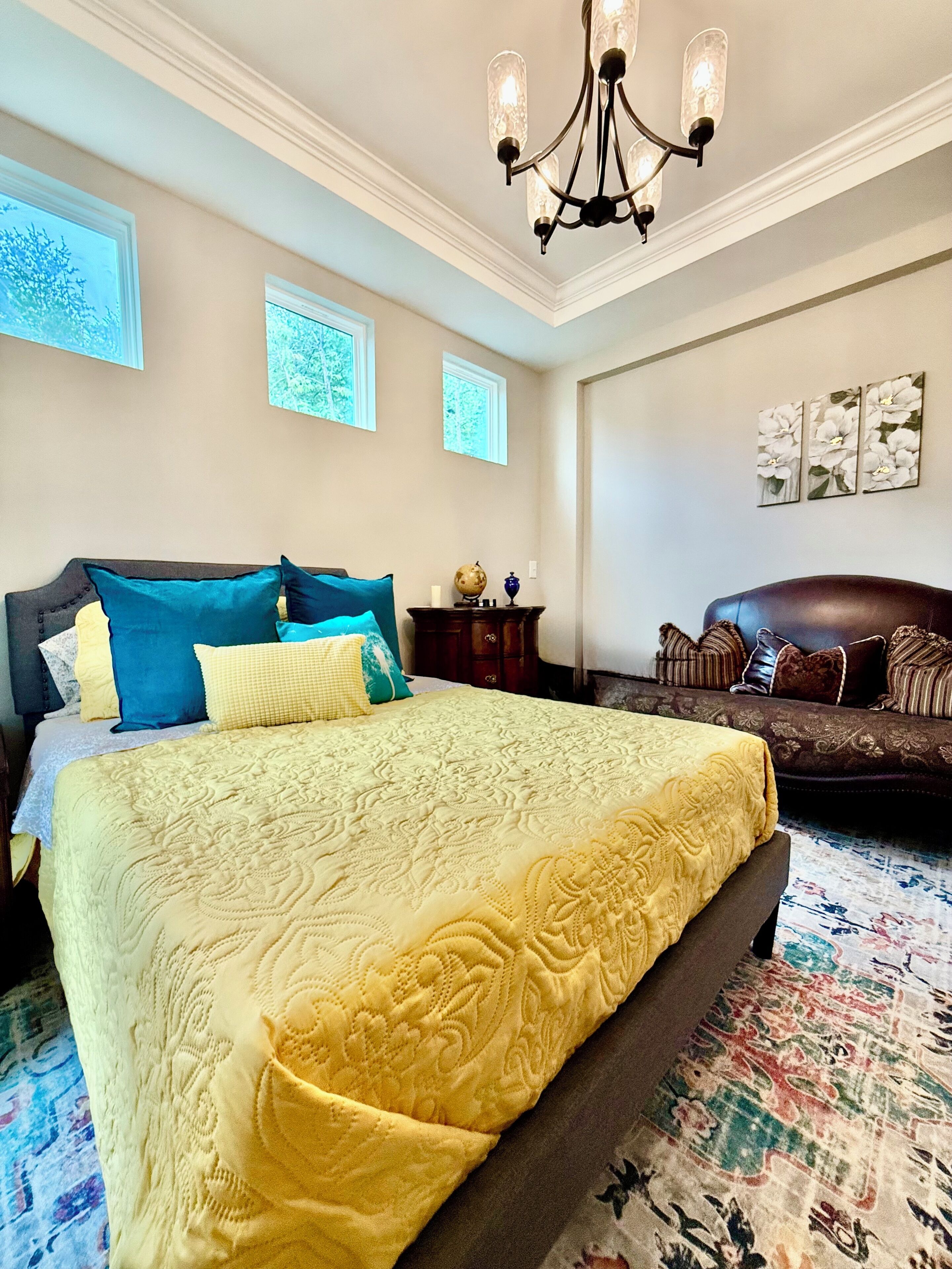 A bedroom with a large bed, a dark sofa, a chandelier, and artwork on the walls.