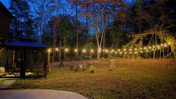 A gazebo with a metal roof and a concrete patio in a backyard with string lights and trees.