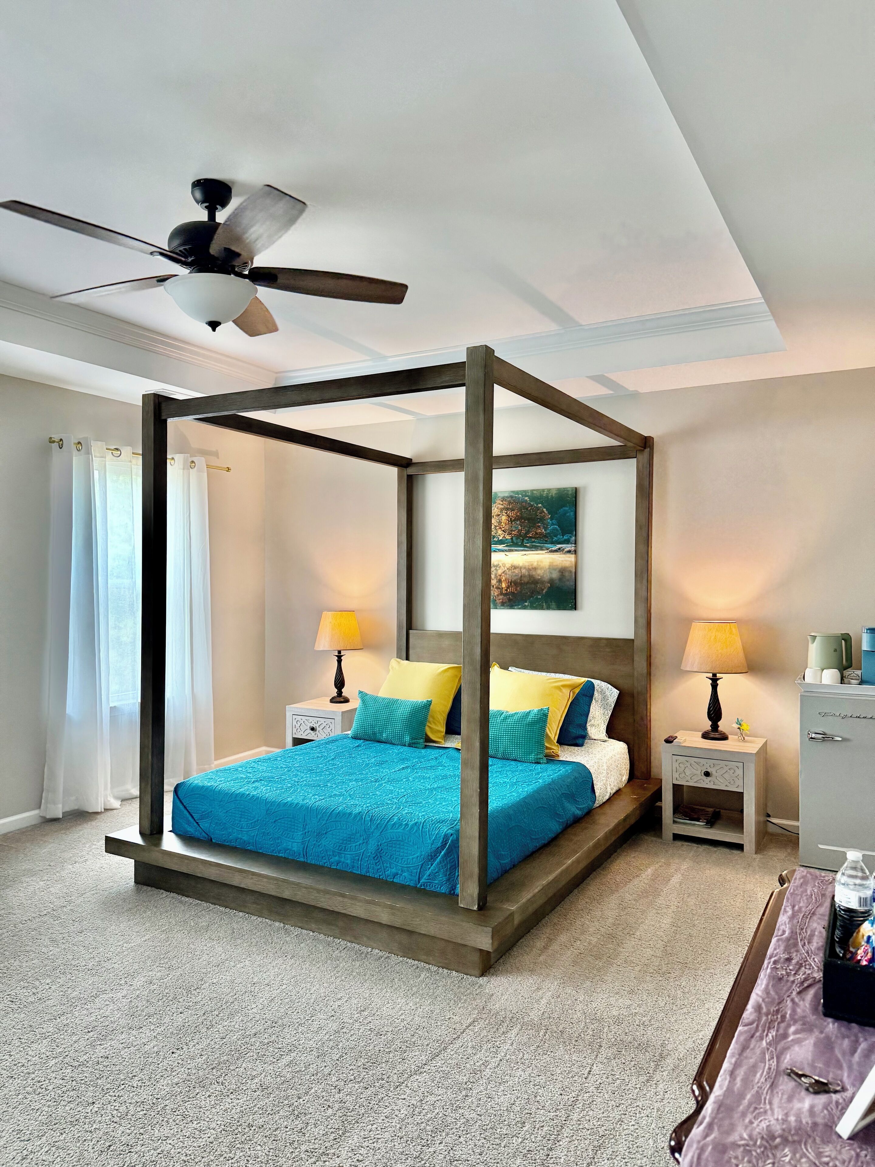 A four-poster bed with a canopy, two bedside tables with lamps, a painting above the bed, and a ceiling fan.