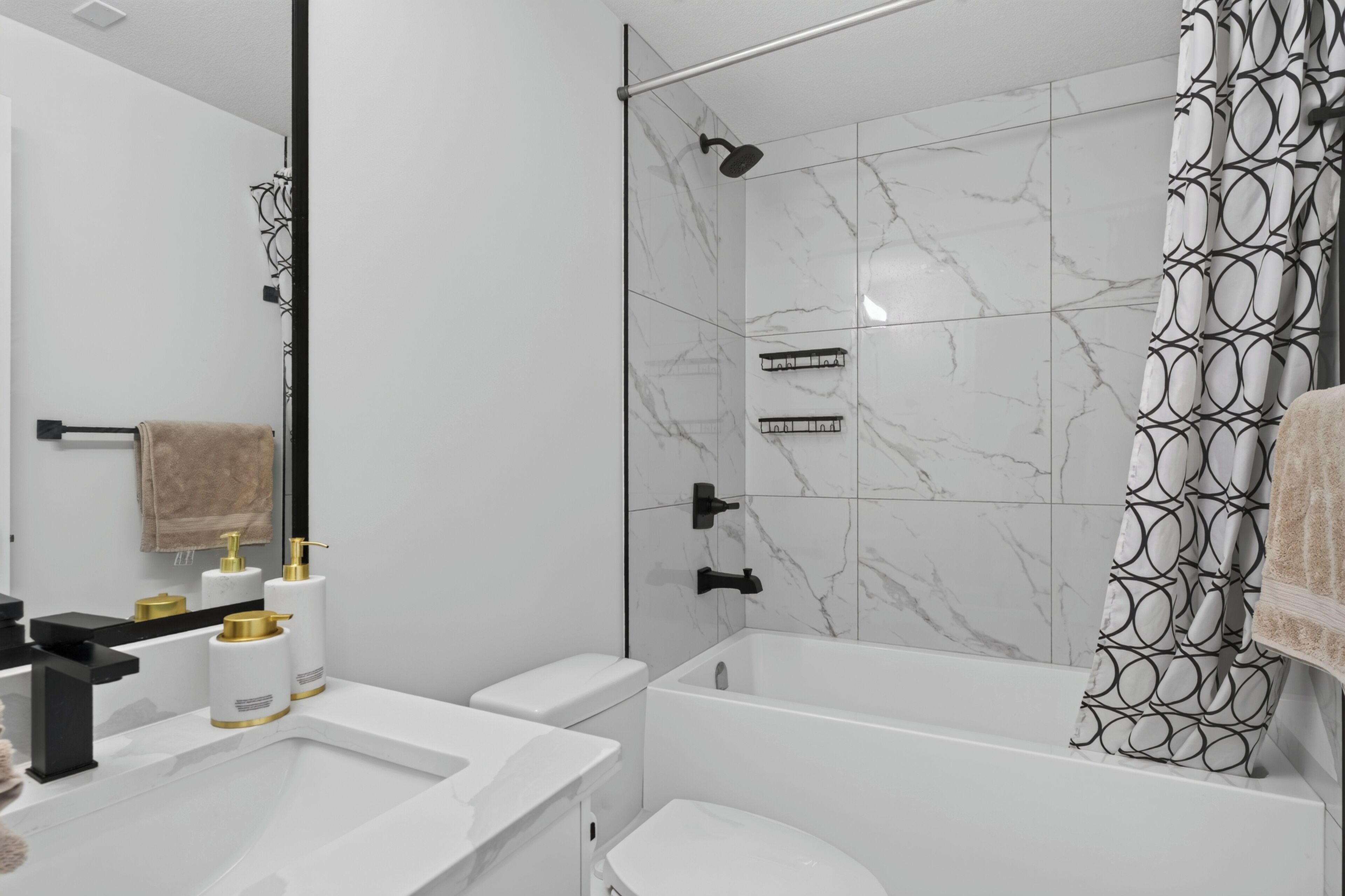 Ein modernes Badezimmer mit einer weißen Marmordusche, einer Badewanne und einer Toilette. Es gibt zwei Waschbecken mit Goldarmaturen sowie einen Handtuchhalter mit einem braunen Handtuch.